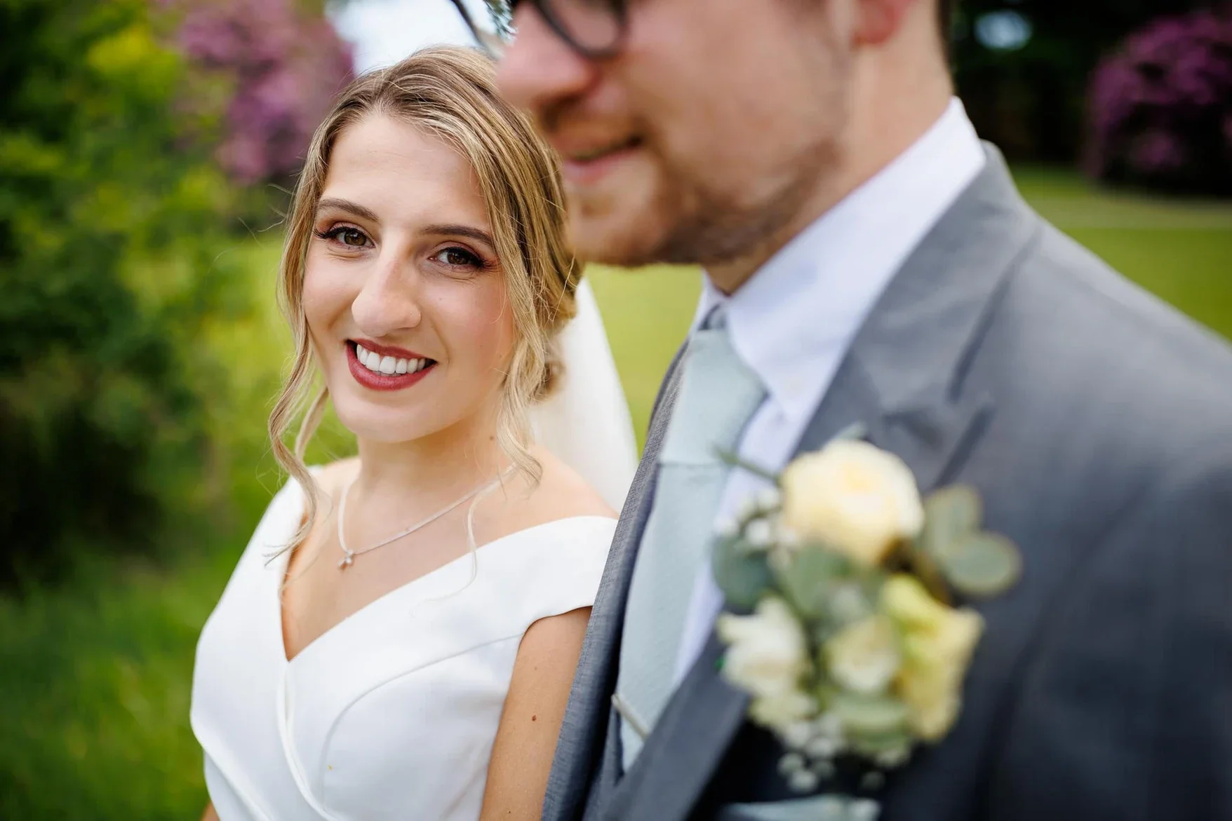 A bride and groom standing close outdoors, with trees and foliage in the background. The bride is smiling at the camera, wearing a white dress and a delicate necklace, with her hair styled in loose waves. The groom, partially visible, wears a gray su
