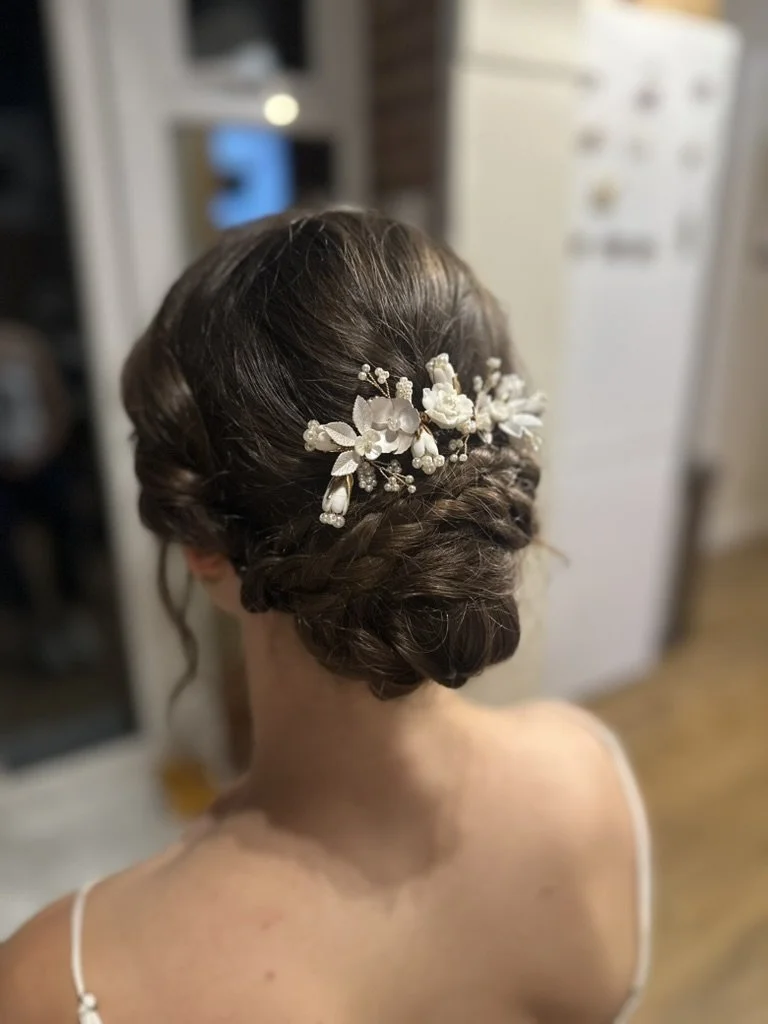 Claire Field Bridal Hair and Make up. Back view of a bride's hairstyle with a braided bun adorned with a white floral hairpiece.