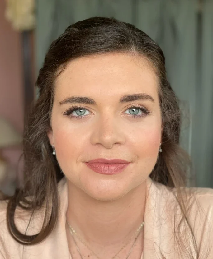 Claire Field Bridal Hair and Make up. Close-up portrait of a woman with light skin, blue eyes, brown hair, and makeup, wearing pearl earrings and layered necklaces, smiling softly. Background is blurred.