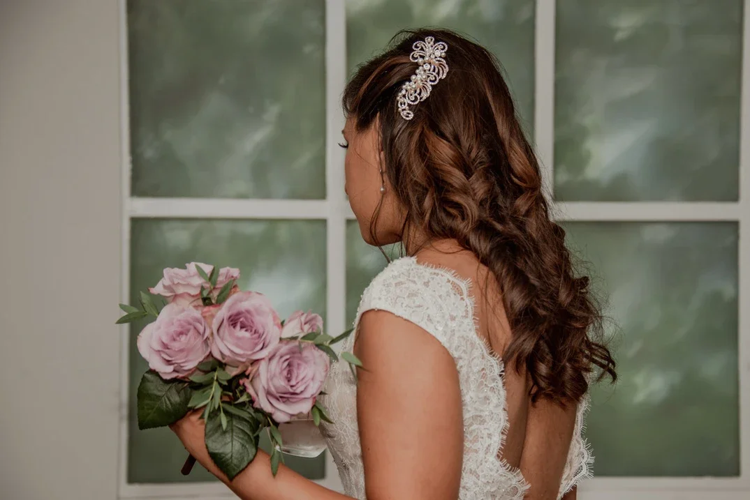 Claire Field Bridal Hair and Make up. A woman wearing a lace dress holding a bouquet of pink roses, with long curly hair and a pearl hair accessory, standing in front of a window with greenery outside.
