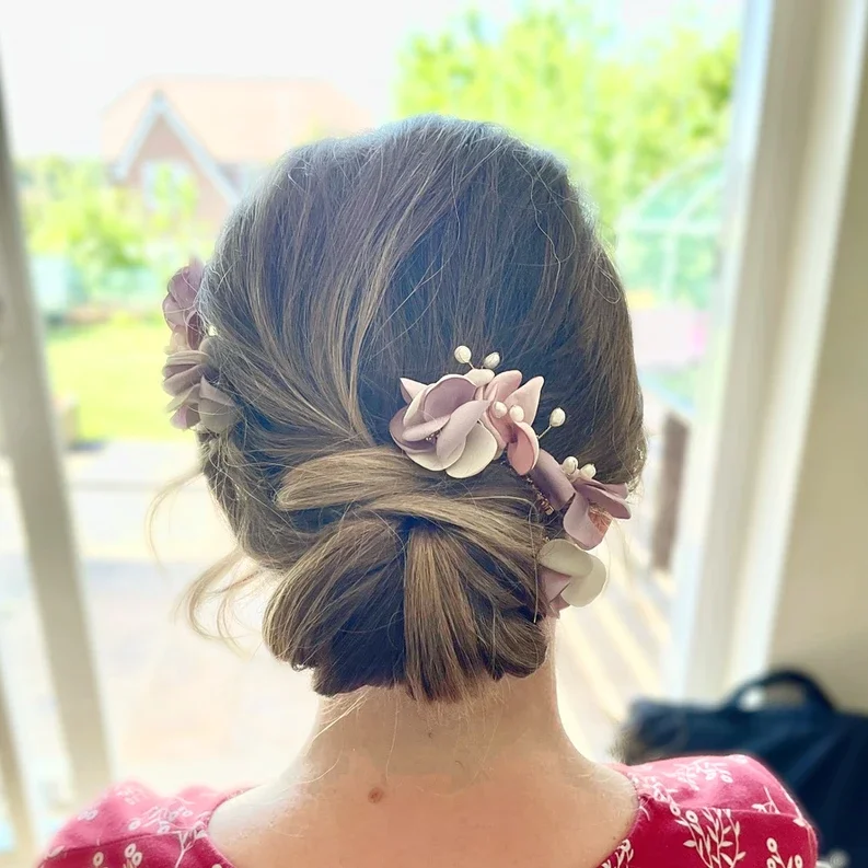 Claire Field Bridal Hair and Make up. A woman with styled hair decorated with pink and purple flowers and pearl accents, standing indoors near a window with a garden view.