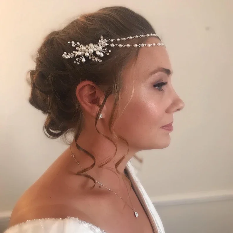 Claire Field Bridal Hair and Make up. A woman with styled hair wearing elegant pearl and crystal hair accessory, pearl earrings, a delicate necklace, and a white garment.