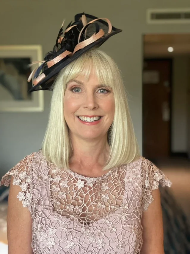 Claire Field Bridal Hair and Make up. A woman with blonde hair wearing a lace dress and a decorative black and pink hat, smiling at the camera.