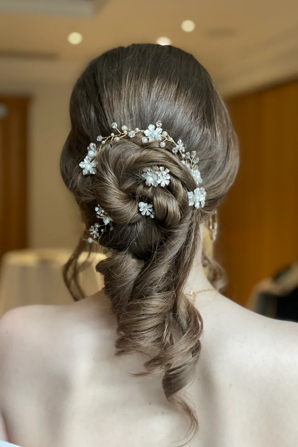 Claire Field Bridal Hair and Make up. Elegant updo hairstyle with braid and loose curls, decorated with floral hair accessories and pearl accents, on a woman with long brown hair.