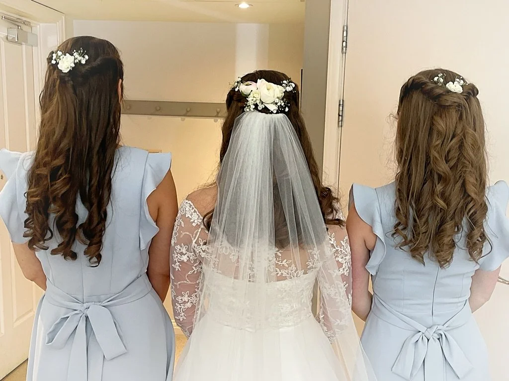 A bride in a lace wedding dress and veil standing between two women in light blue dresses with ruffled sleeves and tied bows, all with floral hair accessories.