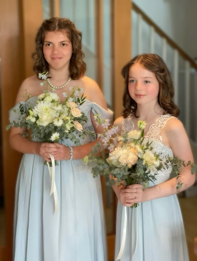 Claire Field Bridal Hair and Make up. Two young girls in matching light blue dresses holding bouquets of white and pastel flowers at a formal event.