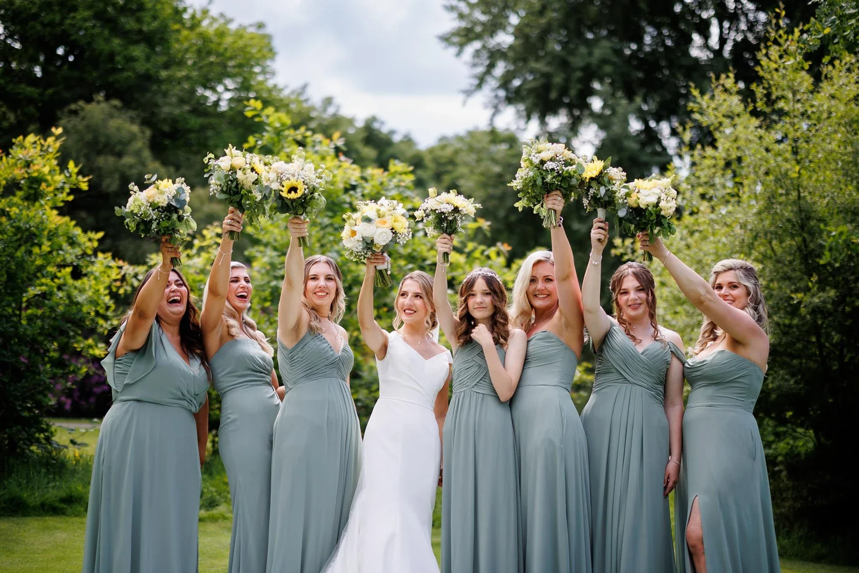 Claire Field Bridal Hair and Make up. A group of seven women, dressed in matching light blue bridesmaid dresses, celebrating outdoors by holding bouquets of white and yellow flowers with sunflowers, on a lush green lawn with trees in the background.