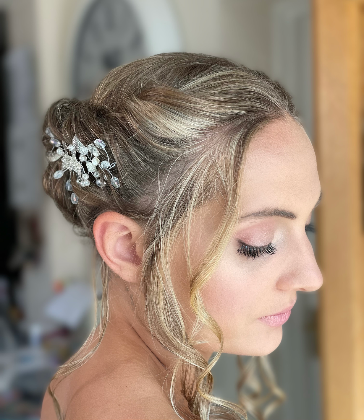 Claire Field Bridal Hair and Make up. Close-up of a bride with styled blond hair, adorned with a decorative hairpiece featuring pearls and crystals, with her eyes closed and makeup.