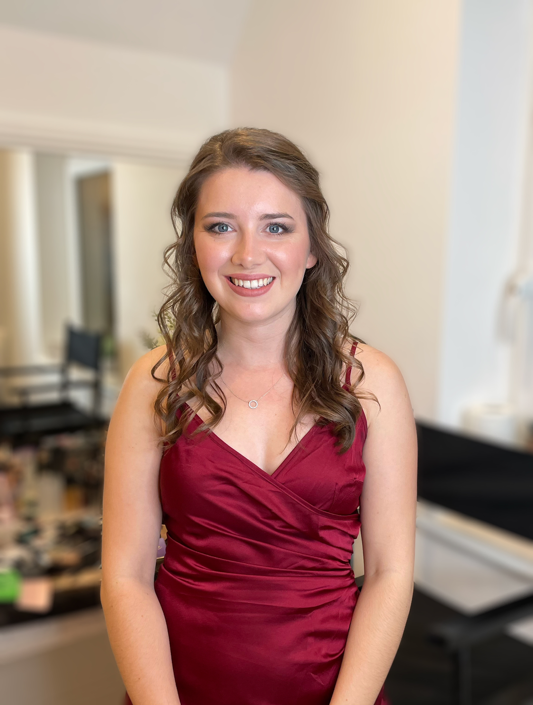 Claire Field Bridal Hair and Make up. A young woman with brown wavy hair and blue eyes wearing a sleeveless burgundy dress, smiling in an indoor setting.