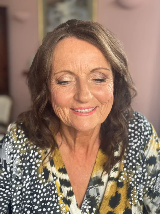 Claire Field Bridal Hair and Make up. Close-up of a smiling middle-aged woman with shoulder-length brown hair wearing a patterned blouse, in a cozy indoor setting.