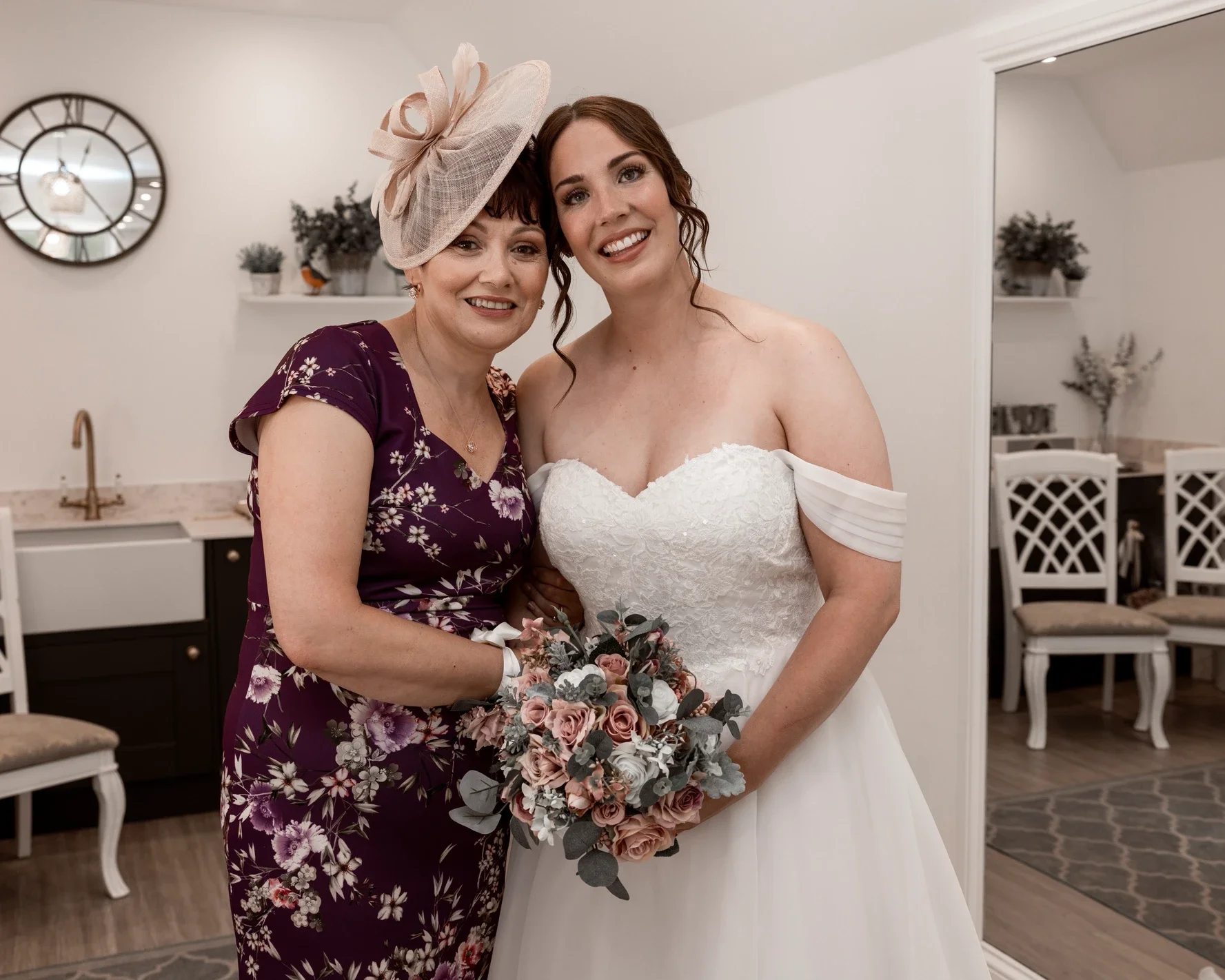 Claire Field Bridal Hair and Make up. The woman in the wedding gown holds a bouquet of pink and grey flowers. The background shows a mirror, a clock, and decorated shelves.