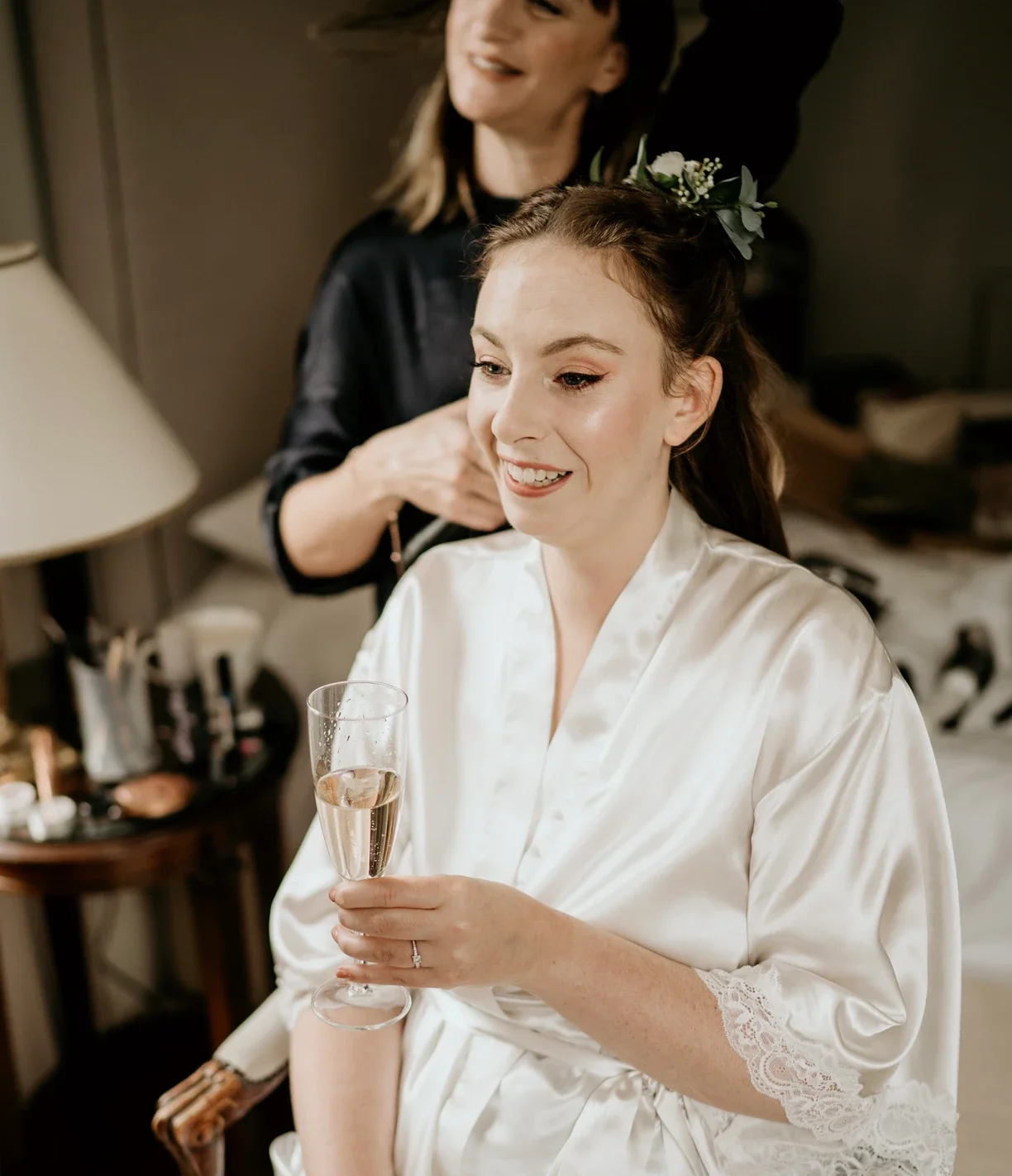 Claire Field Bridal Hair and Make up. A woman in a satin robe smiling while holding a glass of champagne, with another woman helping her with her hair in a room with a lamp and makeup supplies.