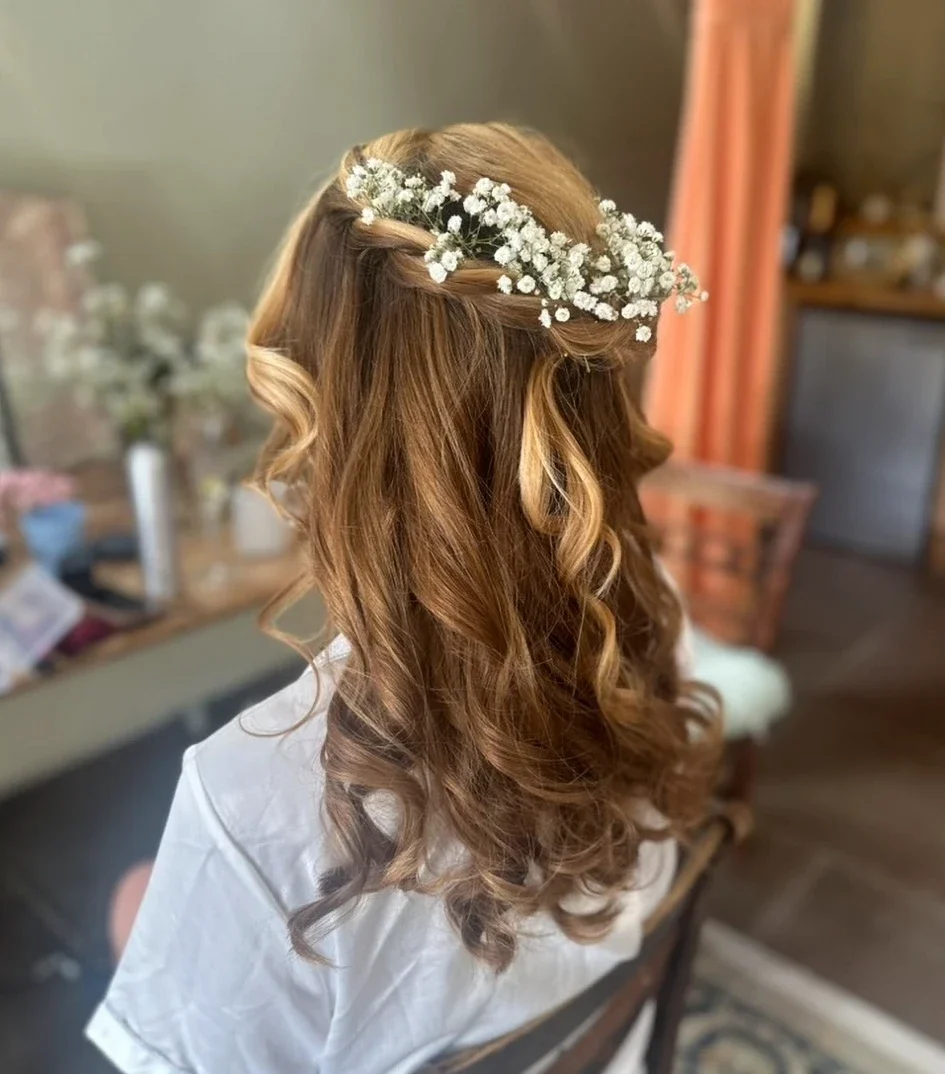 Claire Field Bridal Hair and Make up. Woman with long, wavy red hair wearing a floral headpiece, sitting indoors near a table and chair, with blurred background.