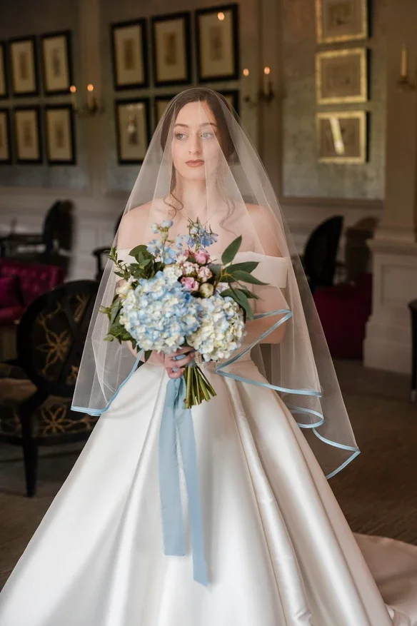 Claire Field Bridal Hair and Make up. Bride in a wedding gown holding a bouquet of pink, blue, and white flowers, standing in an elegant room with framed pictures on the wall.