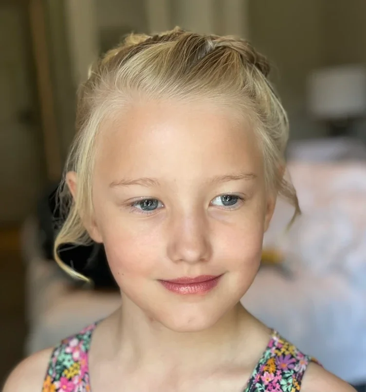 Claire Field Bridal Hair and Make up. Close-up of a young girl with blonde hair styled in a braid, blue eyes, and a gentle smile, wearing a colorful floral top indoors.