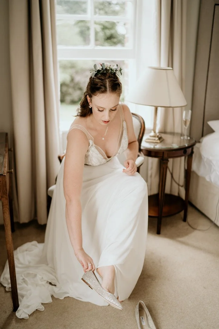 Claire Field Bridal Hair and Make up. A bride in a white wedding dress sitting on a chair, adjusting her silver shoe in a warmly lit room with beige curtains and a bedside lamp.
