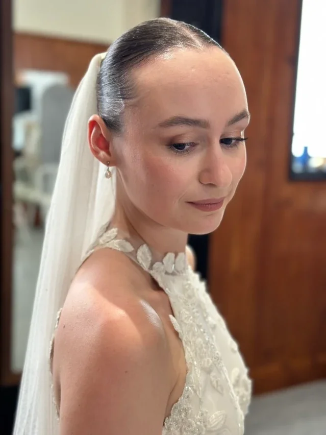 Claire Field Bridal Hair and Make up. A bride with soft makeup, wearing a lace wedding dress with floral appliqué, and a veil attached to her hair, standing indoors.