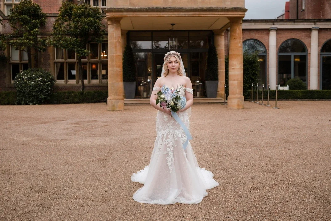 Claire Field Bridal Hair and Make up. Bride in a white lace wedding gown holding a bouquet of flowers standing outside a brick building with large windows and trees.