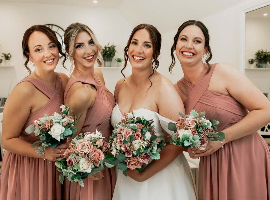 Claire Field Bridal Hair and Make up. Four women in pink bridesmaid dresses and one bride in white dress smiling and holding bouquets of flowers at a wedding.