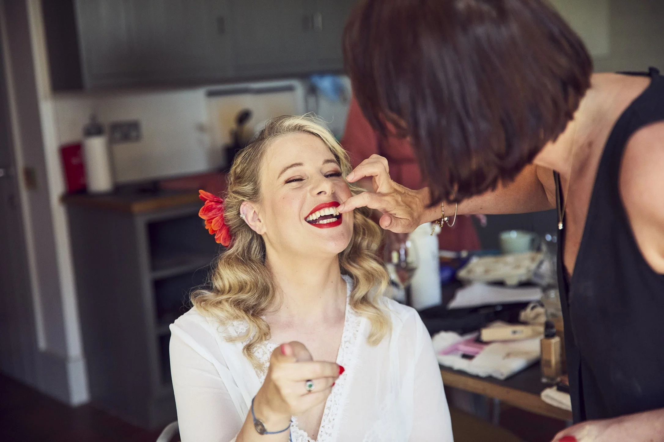 Claire Field Bridal Hair and Make up. A woman with blonde curly hair, red lipstick, and a flower in her hair is smiling and looking at a makeup artist applying makeup on her face in a room with kitchen cabinets in the background.