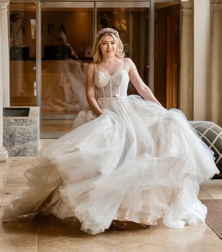 Claire Field Bridal Hair and Make up. A woman in a wedding gown sitting inside a building with glass doors, with her reflection visible in the glass behind her.