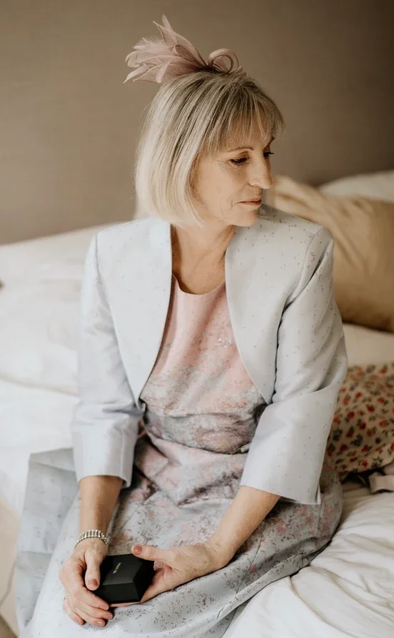 Claire Field Bridal Hair and Make up. A woman sitting on a bed, looking to her right, holding a small black box in her hands, wearing a white blazer and a pastel dress, with a pink fascinator in her blonde bob haircut.