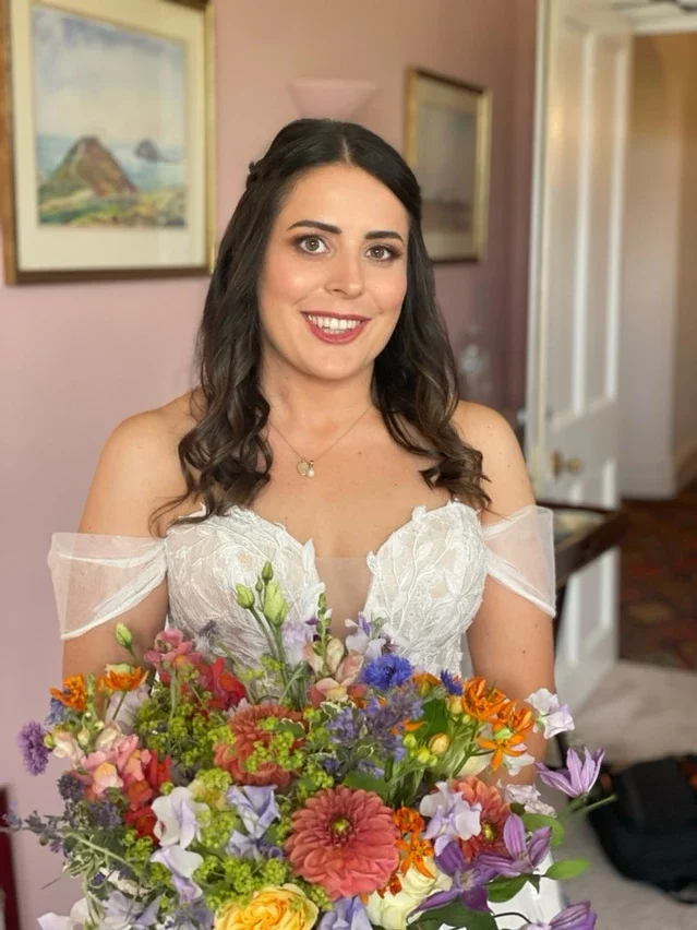 Claire Field Bridal Hair and Make up. A woman in a white lace dress holding a large colorful bouquet of flowers, smiling indoors with framed artwork on the wall behind her.