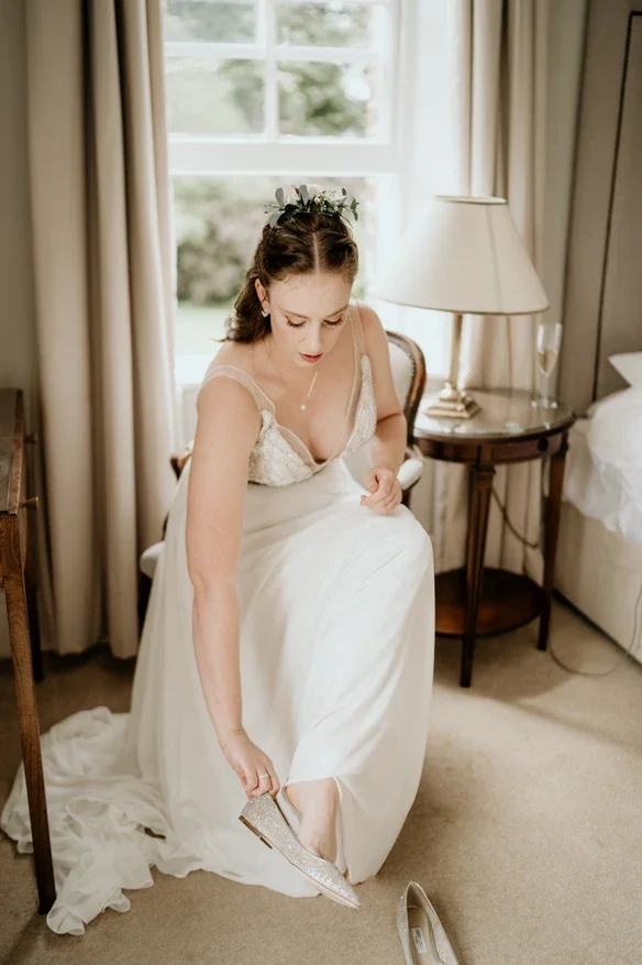 Claire Field Bridal Hair and Make up. A bride sitting on a chair in a hotel room, putting on her sparkly shoe, with a window and curtains behind her and a side table with a lamp and a glass of champagne.