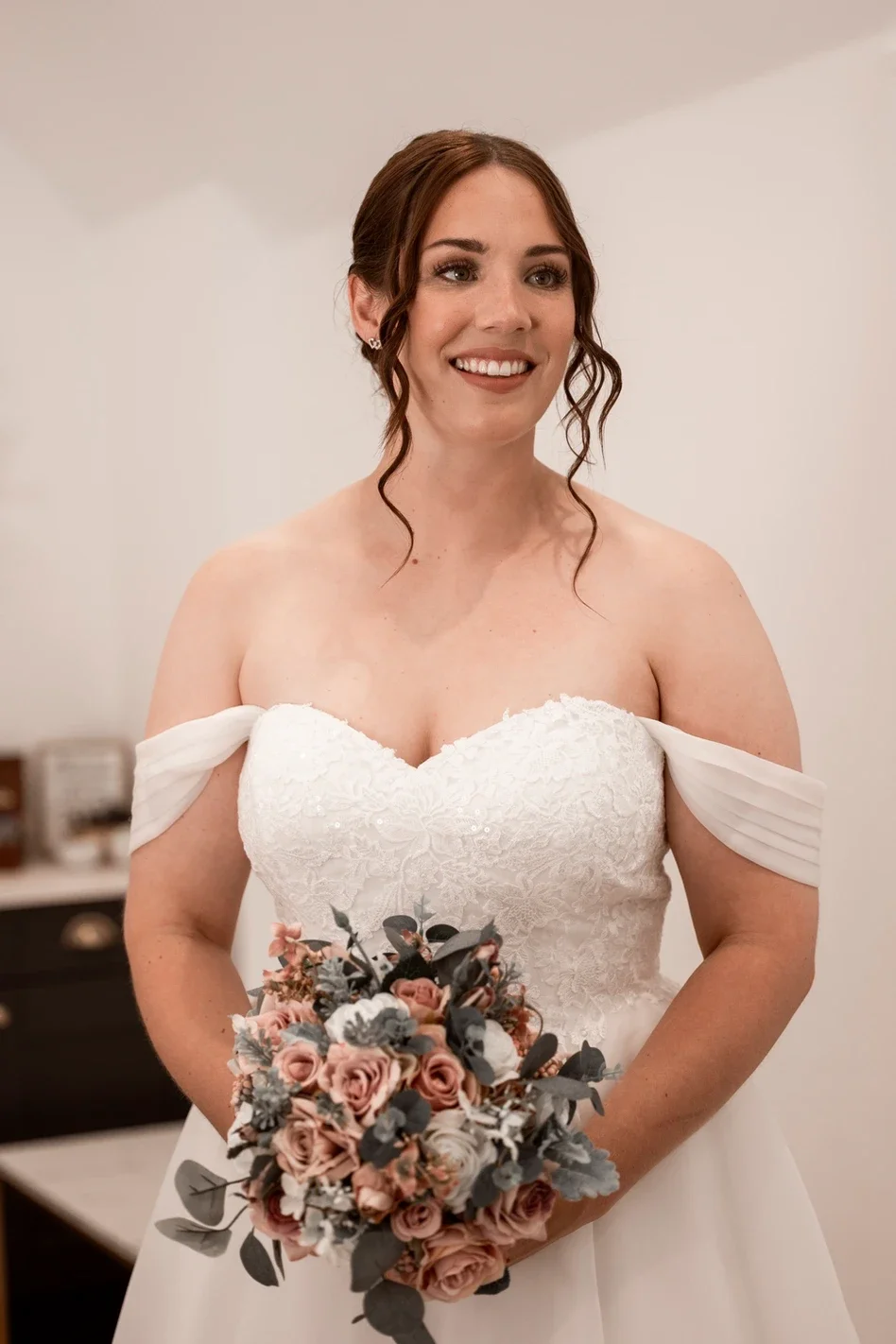 Claire Field Bridal Hair and Make up. A smiling bride with red hair, wearing a strapless white lace wedding gown, holding a bouquet of pink and gray flowers, standing indoors against a plain background.