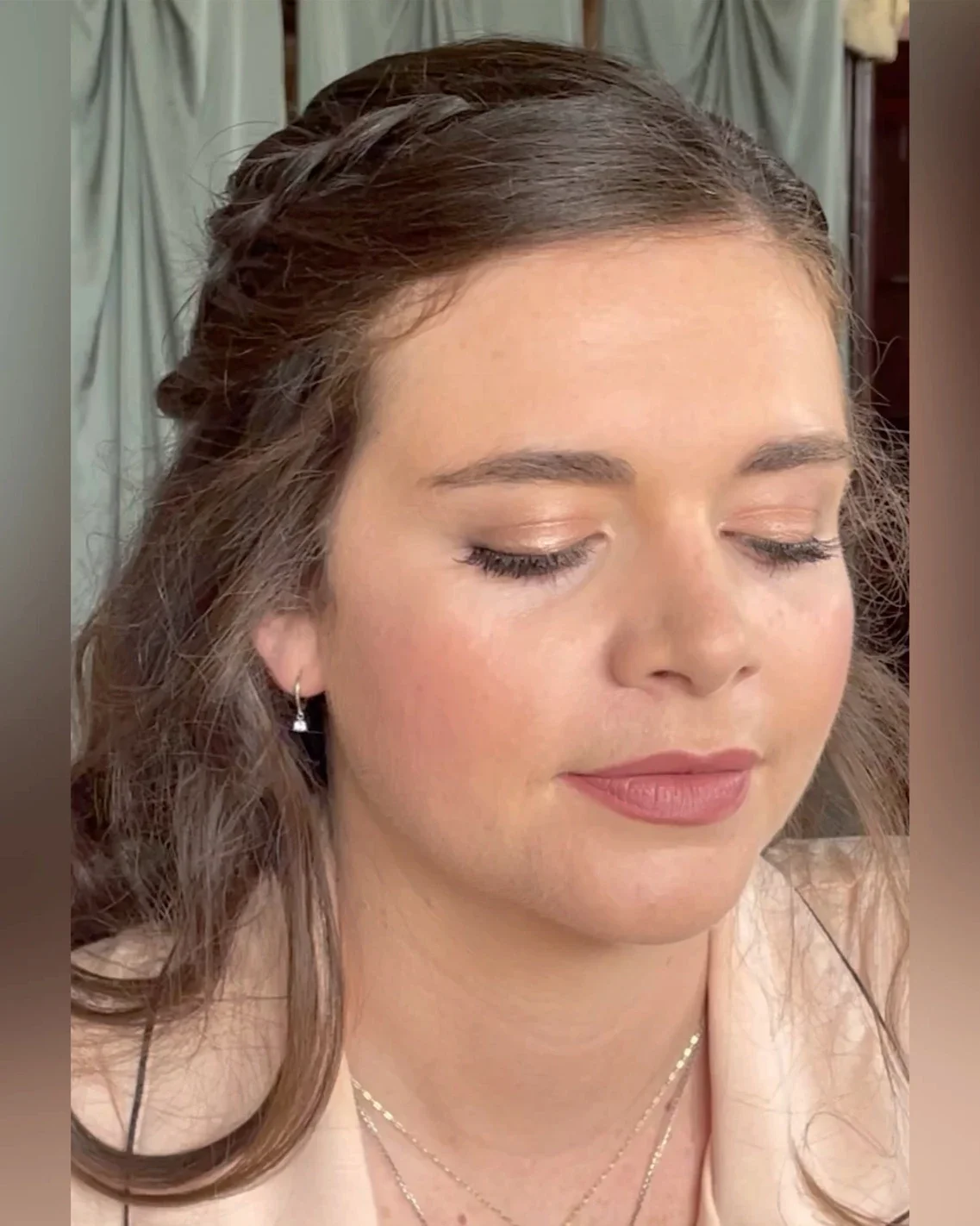 Claire Field Bridal Hair and Make up. A close-up of a woman with brown hair and fair skin with her eyes closed. She is wearing earrings and layered necklaces, and the background shows a curtain.