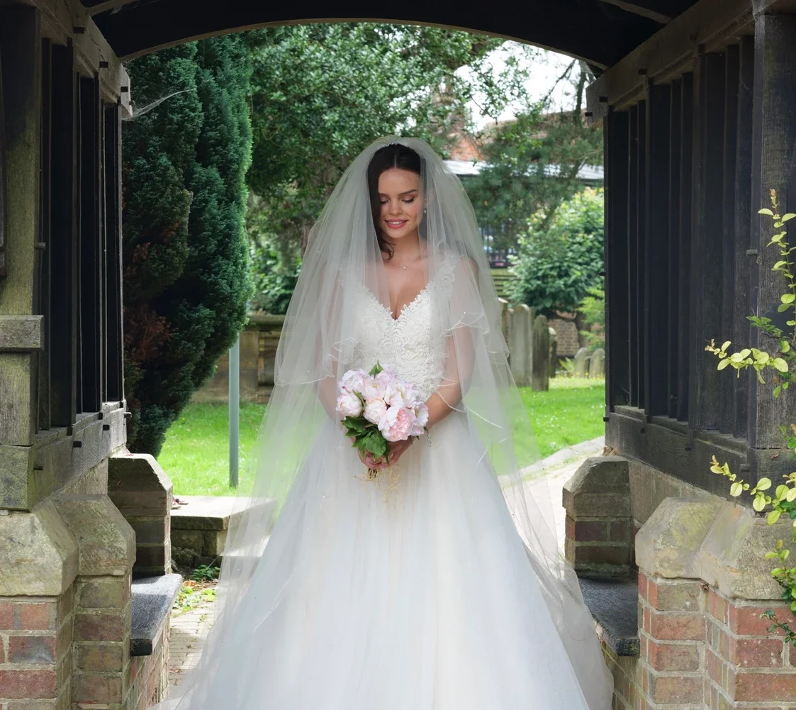 Claire Field Bridal Hair and Make up. A bride in a white wedding gown and veil holding a bouquet of pink flowers standing under a stone and black metal archway in a garden.