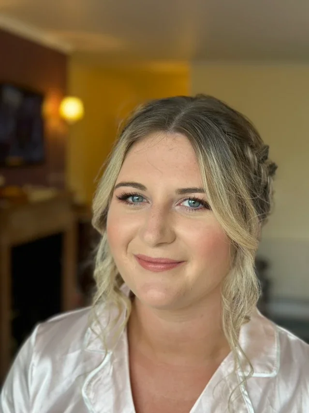 Claire Field Bridal Hair and Make up. Portrait of a woman with blonde hair and blue eyes, wearing a white satin robe, smiling in a warmly lit room.