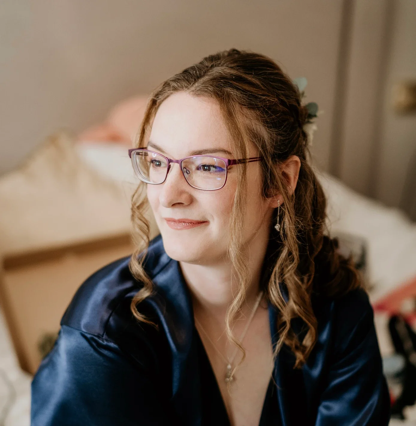Claire Field Bridal Hair and Make up. A woman with light brown curly hair and glasses, smiling softly, wearing a dark satin robe, sitting in a warmly lit room.