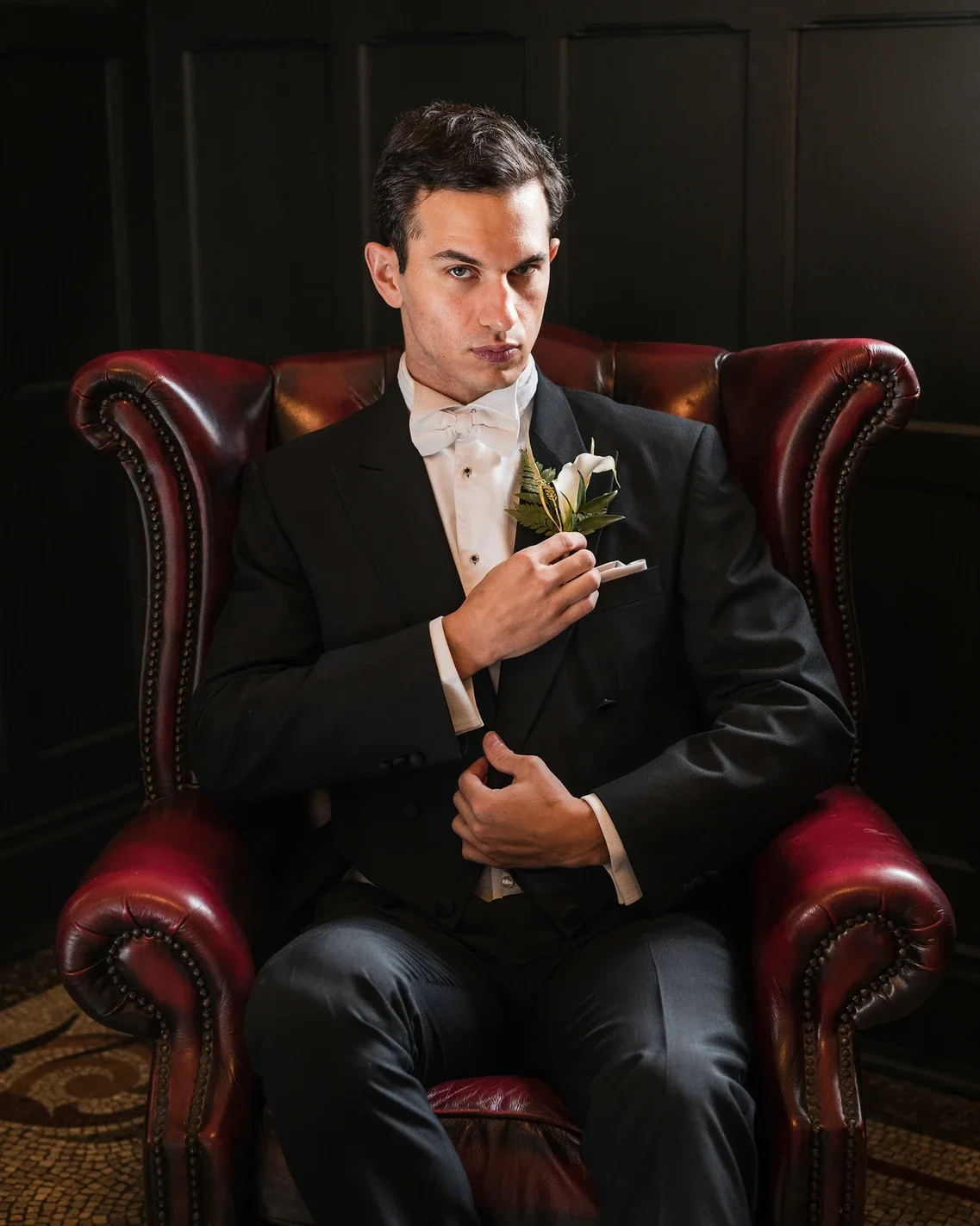 Claire Field Bridal Hair and Make up. A young man dressed in a black tuxedo with a white bow tie and boutonniere, sitting in a red leather armchair with ornate wooden details.