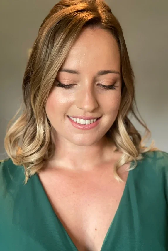 Claire Field Bridal Hair and Make up. A smiling woman with wavy blonde hair and fair skin, wearing a green top with a V-neckline.