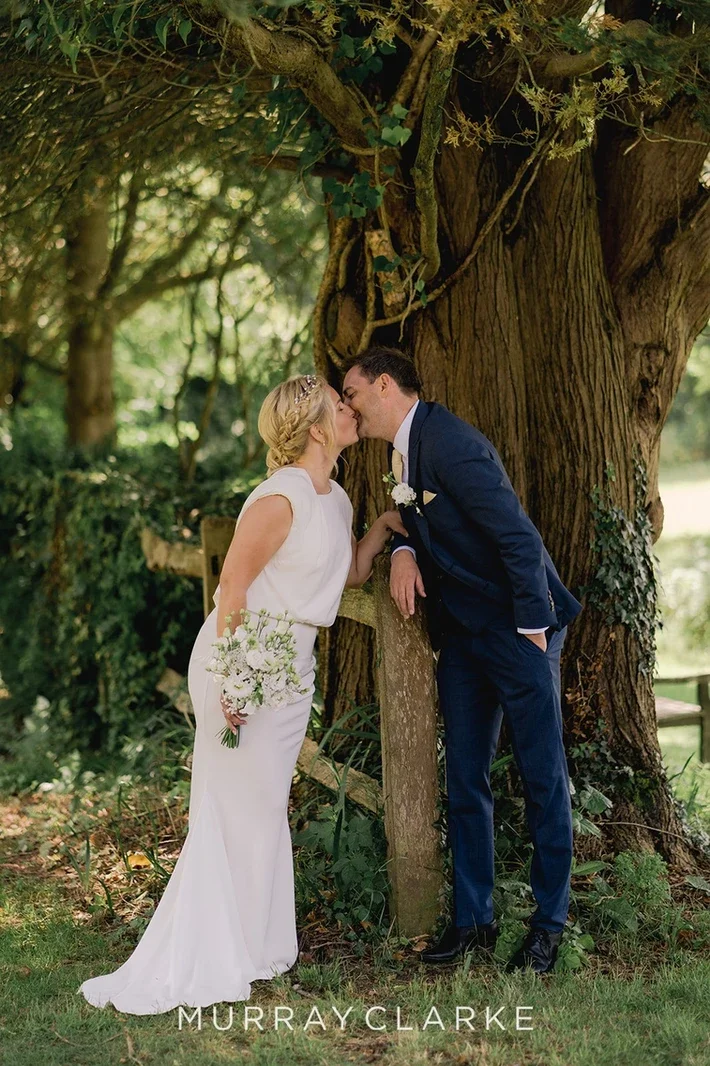 Claire Field Bridal Hair and Make up. A bride and groom sharing a kiss outdoors in a wooded area during their wedding, with a large tree in the background. The bride is holding a bouquet of white flowers, and the groom is dressed in a dark blue suit.