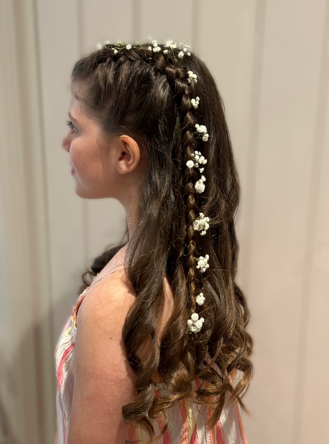 Claire Field Bridal Hair and Make up. A young girl with long wavy brown hair decorated with small white flowers, styled in braids, with a side profile view.