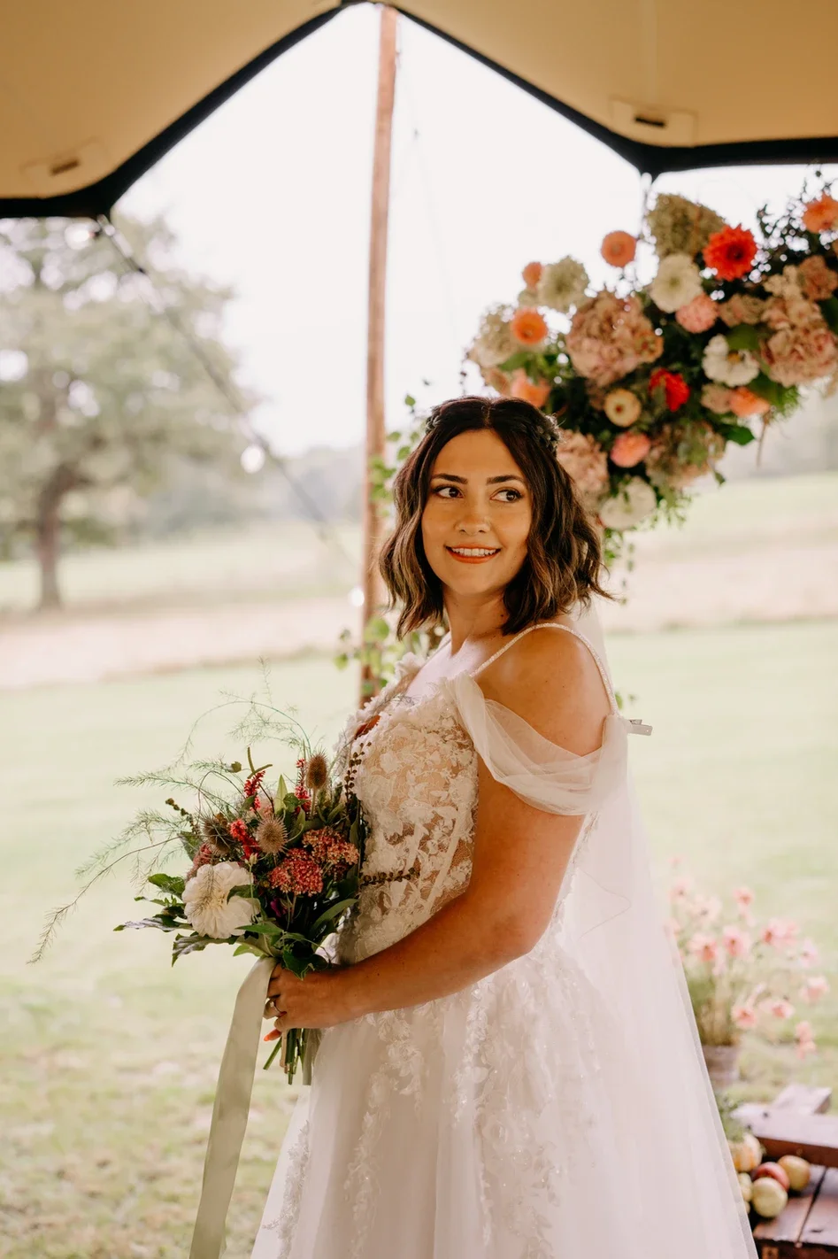 Claire Field Bridal Hair and Make up. A bride in a white lace wedding dress holding a bouquet of flowers, smiling and posing outdoors under a tent with a large flower arrangement overhead.