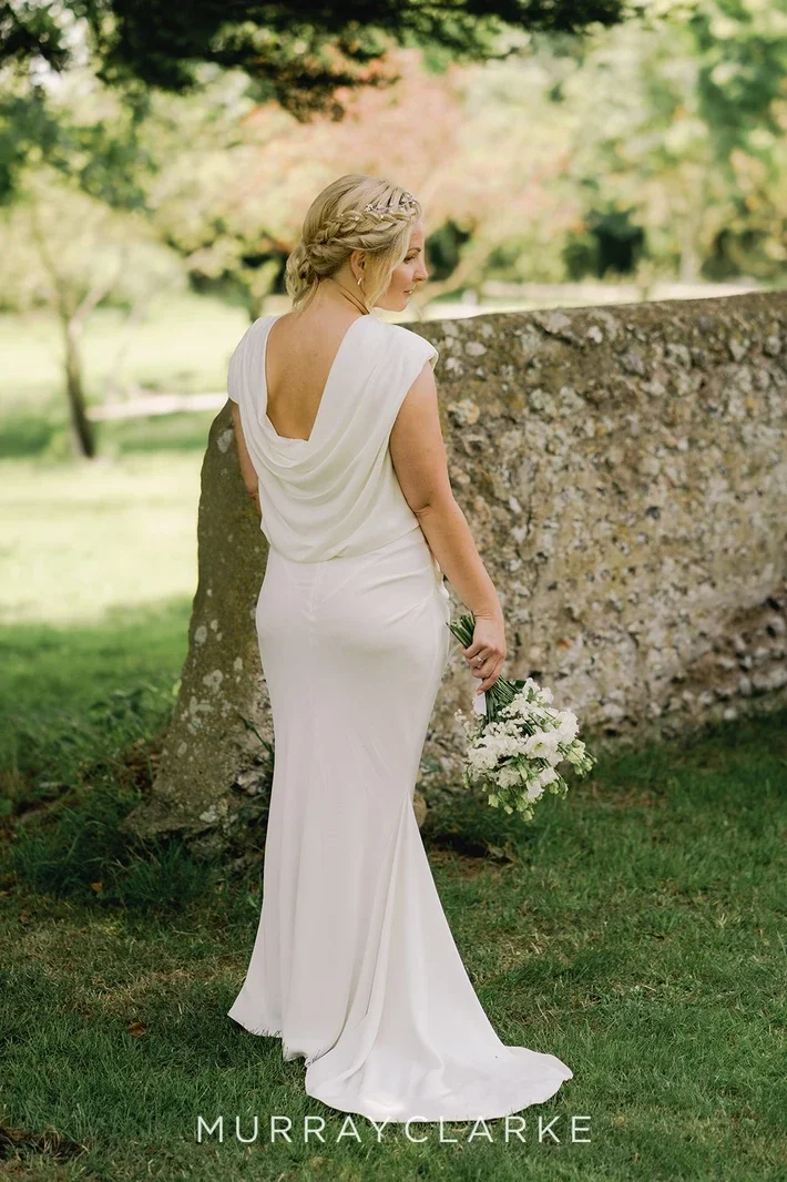 Claire Field Bridal Hair and Make up. A woman in a white gown holding a bouquet of white flowers, standing outdoors near a stone wall, with trees and greenery in the background.