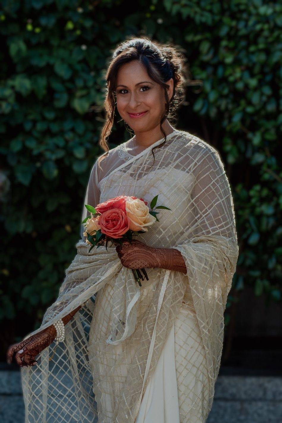 Claire Field Bridal Hair and Make up. A woman in a cream-colored saree holding a bouquet of roses, standing outdoors with greenery in the background, smiling at the camera.