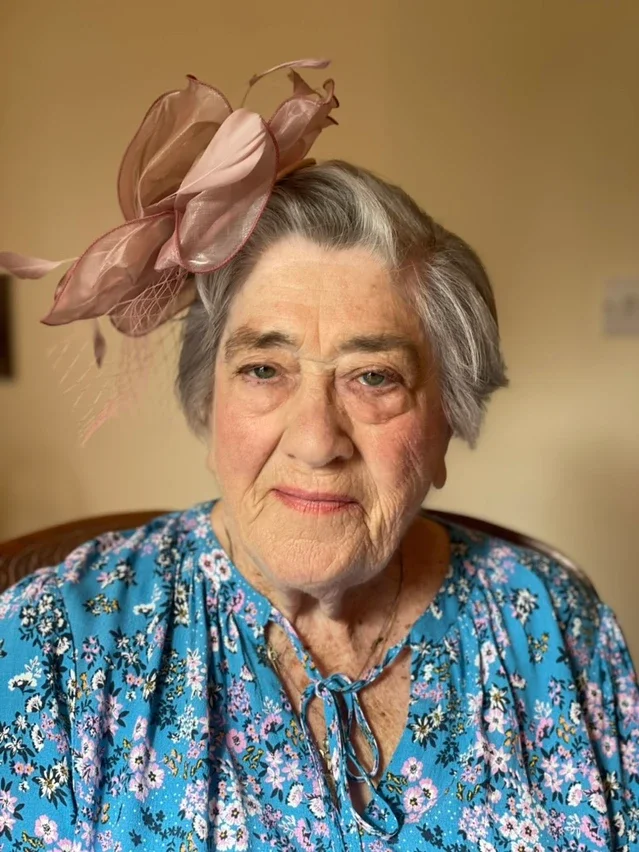 Claire Field Bridal Hair and Make up. An elderly woman with grey hair wearing a large pink floral hat and a blue floral blouse.