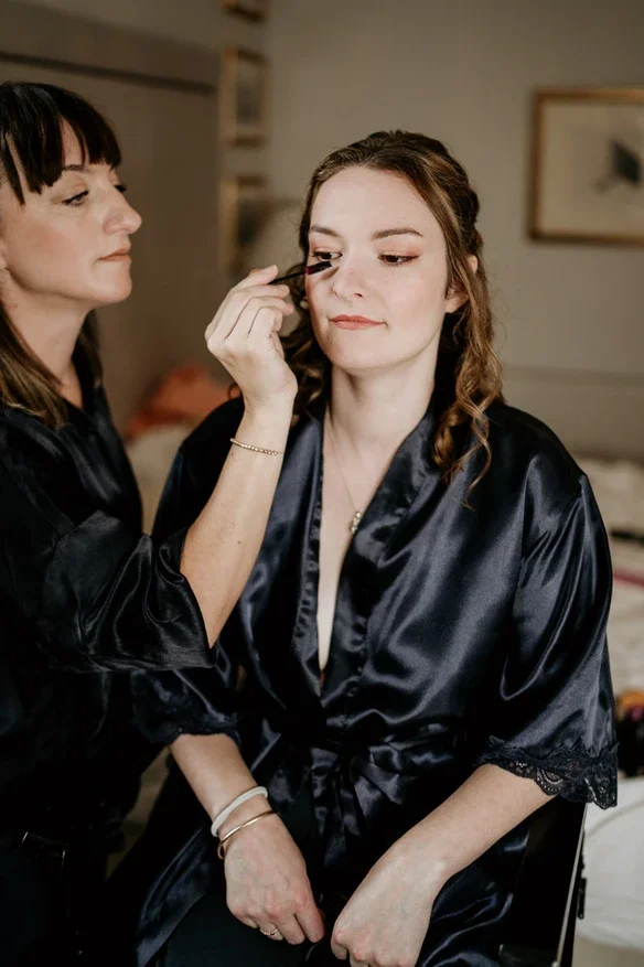 Claire Field Bridal Hair and Make up. A woman is applying makeup to another woman who is sitting. Both are dressed in black robes, likely preparing for a special event.