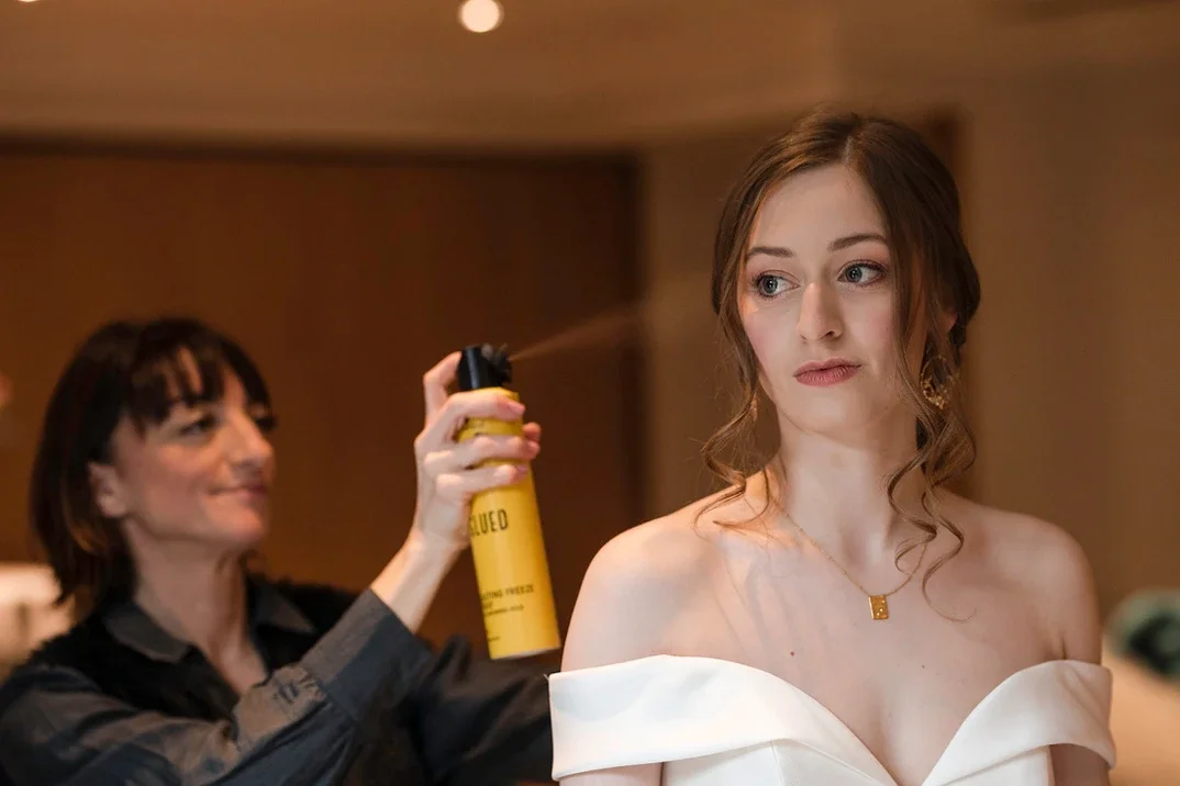 Claire Field Bridal Hair and Make up. A woman with wavy brown hair wearing a white off-the-shoulder dress sits while a hairstylist sprays hairspray on her hair in a salon or dressing room.