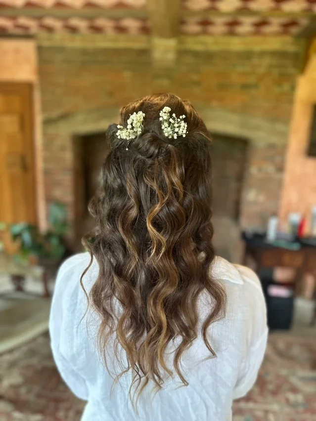 Claire Field Bridal Hair and Make up. Back view of a woman with long, wavy hair styled in loose curls, adorned with a small bouquet of white flowers on top.