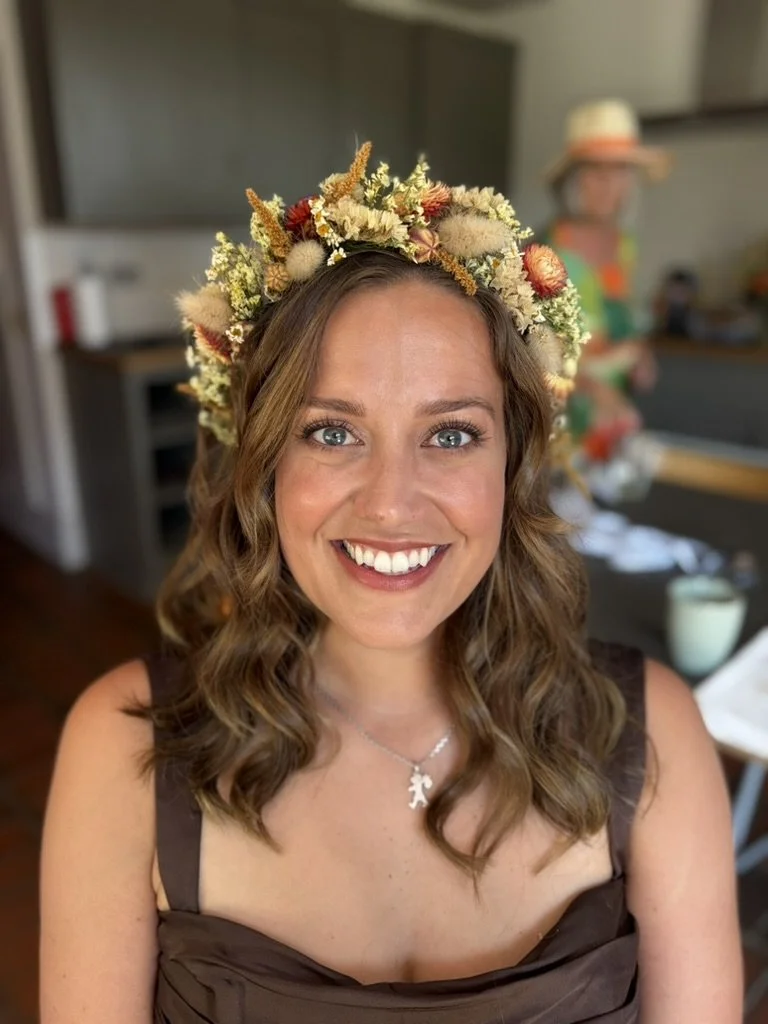 A young woman with wavy brown hair and blue eyes, wearing a floral crown and a brown dress, smiling in an indoor setting.