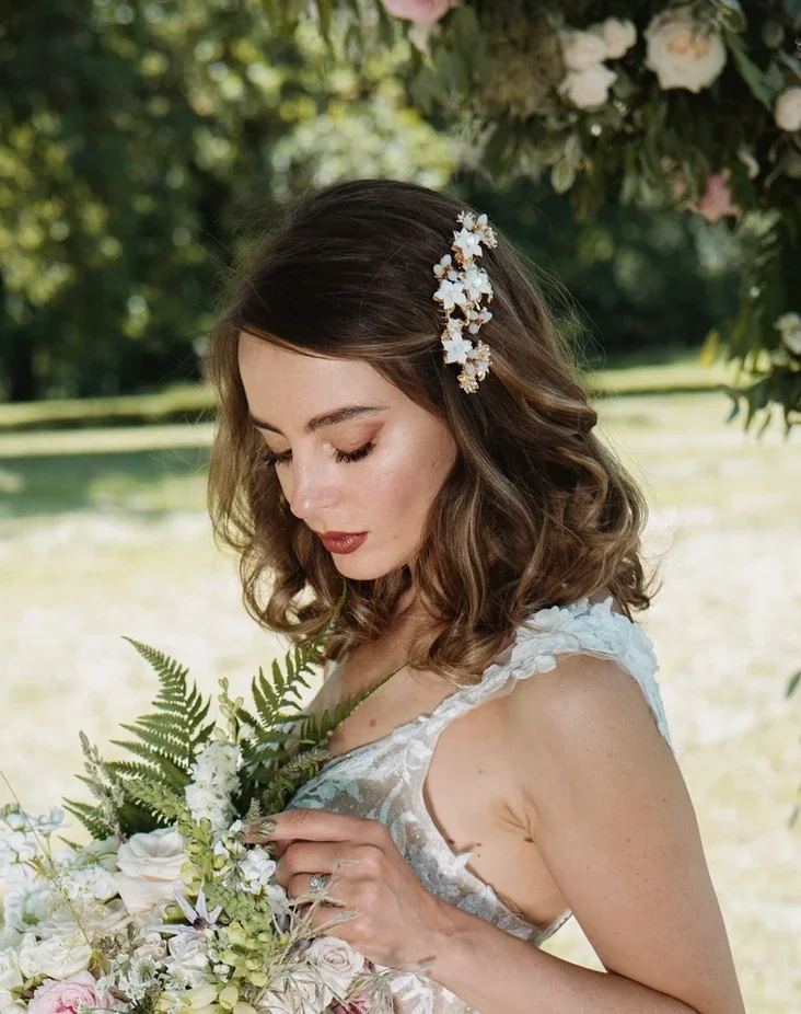Claire Field Bridal Hair and Make up. A woman with shoulder-length wavy brown hair, wearing a white floral hair accessory, holding a bouquet of flowers outdoors under a tree, with greenery in the background.