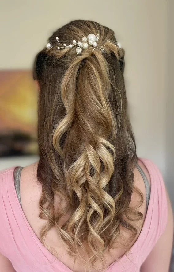 Claire Field Bridal Hair and Make up. Back view of a woman with styled medium-length, wavy, blonde hair adorned with a pearl and bead hair accessory, wearing a pink top.