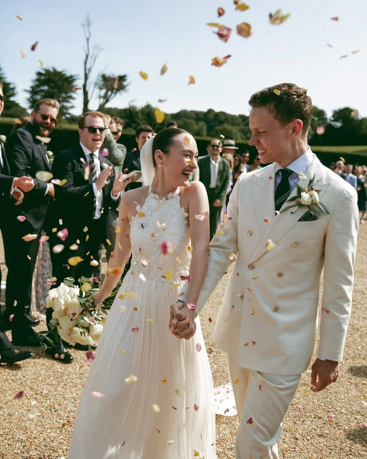 Claire Field Bridal Hair and Make up. A bride and groom hold hands and smile at each other during their wedding celebration, with friends and family celebrating and throwing flower petals in the air.