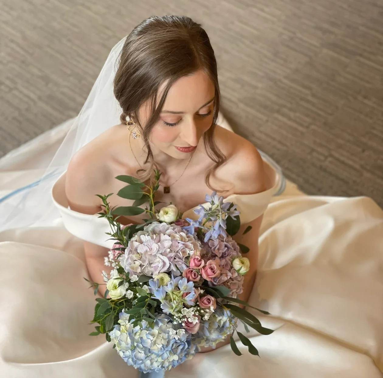 Claire Field Bridal Hair and Make up. A bride in a wedding dress holding a bouquet of pastel-colored flowers, looking down.