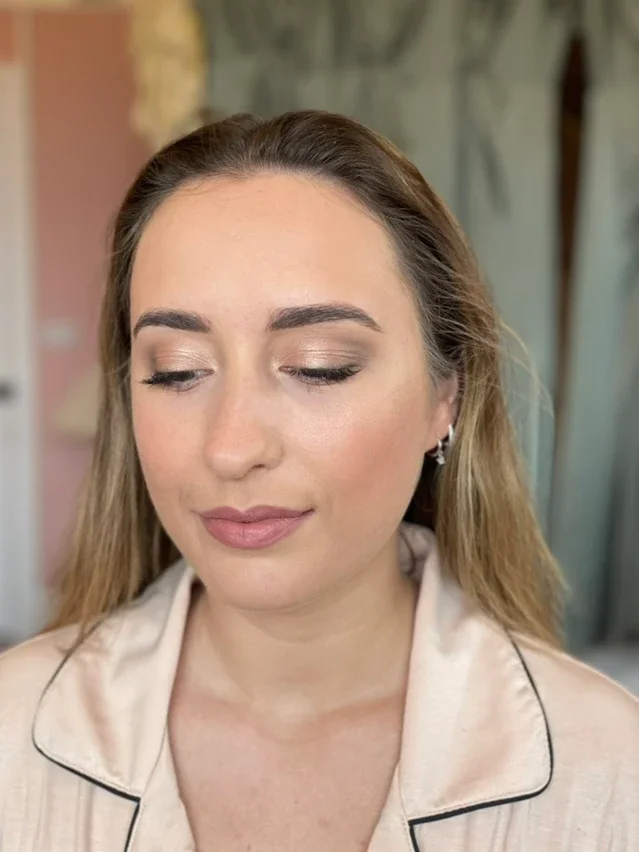 Claire Field Bridal Hair and Make up. Close-up of a woman with light makeup and closed eyes, wearing a beige satin or silk pajama top with black piping, earrings, and long hair.